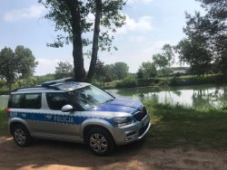 Na zdjęciu widać oznakowany radiowóz policyjny marki skoda yeti, który jest zaparkowany w rejonie akwenu wodnego. W tel widać drzewa i krzewy
